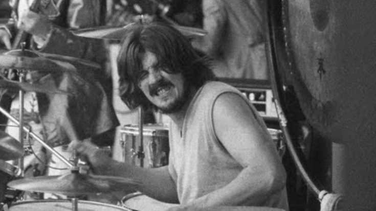 John Bonham intensely playing drums during a live outdoor Led Zeppelin performance, wearing a sleeveless shirt and surrounded by cymbals and percussion equipment.