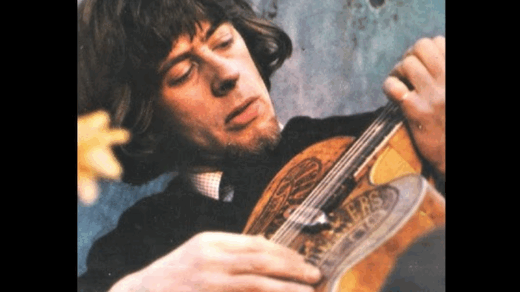 John Mayall sits calmly in a sunlit room, wearing a patterned short-sleeve shirt with his hands gently resting on his lap, surrounded by musical instruments and warm wooden tones.