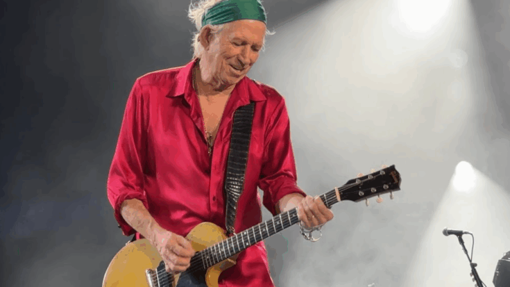 Keith Richards playing guitar on stage wearing a red shirt and green headband under bright stage lights