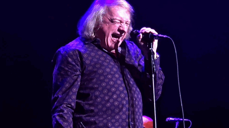 Lou Gramm passionately singing into a microphone during a live stage performance under purple and blue lighting