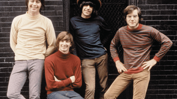 The Lovin' Spoonful band members pose casually against a dark brick wall, each wearing colorful 1960s-style sweaters and smiling at the camera.