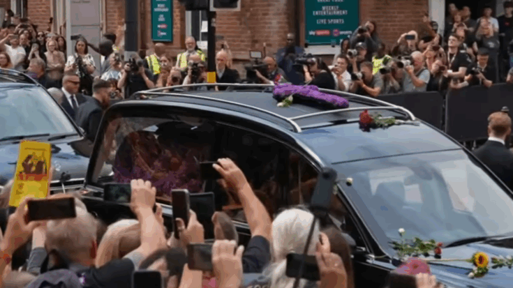 A large crowd gathers in Birmingham as Ozzy Osbourne’s funeral hearse passes through Broad Street, with fans holding up phones and flowers placed on the vehicle.