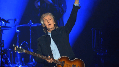 Paul McCartney raises his fist while performing on stage with his iconic Höfner bass guitar, lit by deep blue stage lights.