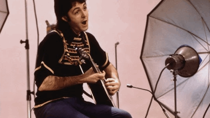 Paul McCartney seated in a studio setting, wearing a black and gold military-style jacket while playing an acoustic guitar and singing, with large photography lights surrounding him.