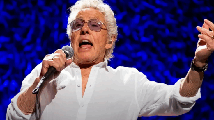 Roger Daltrey passionately singing on stage with a microphone in hand, wearing a white shirt and sunglasses against a vivid blue textured background.