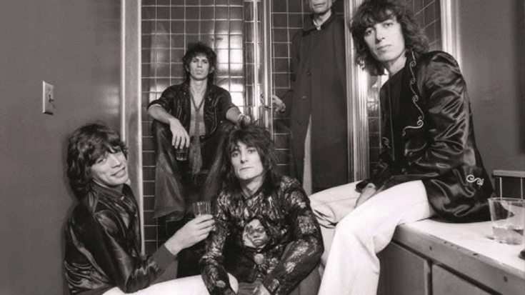 The Rolling Stones relaxing backstage in a tiled room during the late 1970s, with Mick Jagger smiling at the front and Ronnie Wood seated at the center holding a drink.