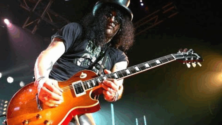 Slash performing live on stage with his signature Les Paul guitar and top hat under concert lights