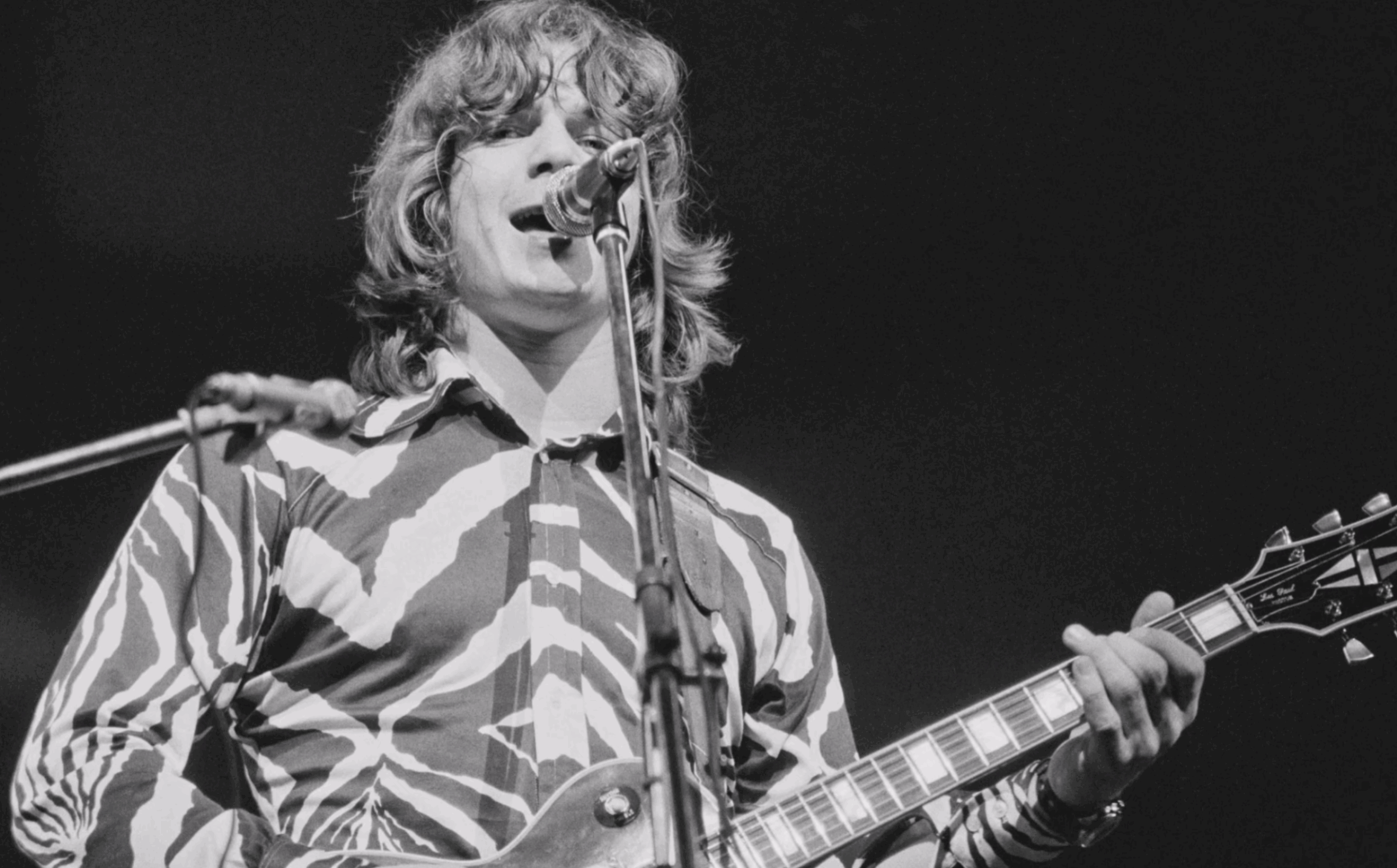 Steve Miller performing live in the 1970s, wearing a patterned shirt and playing a Gibson Les Paul guitar while singing into a microphone.
