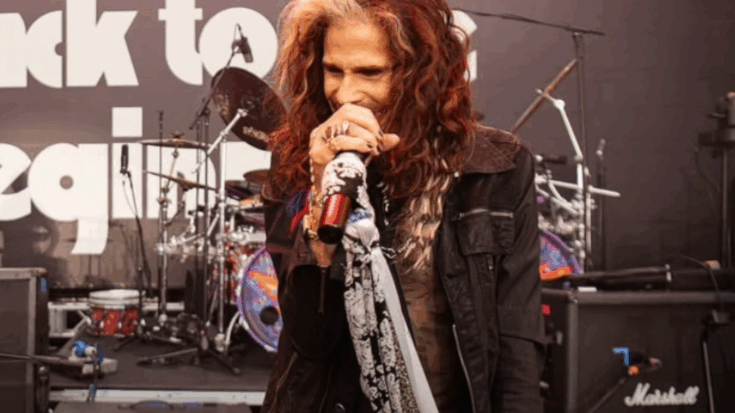 Steven Tyler singing into a microphone on stage, wearing a black jacket and holding a mic wrapped in scarves, with a drum set and Marshall amps behind him.