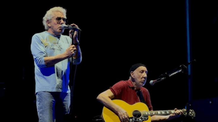 Roger Daltrey sings into a microphone while Pete Townshend plays acoustic guitar during a live performance by The Who on a dark stage.