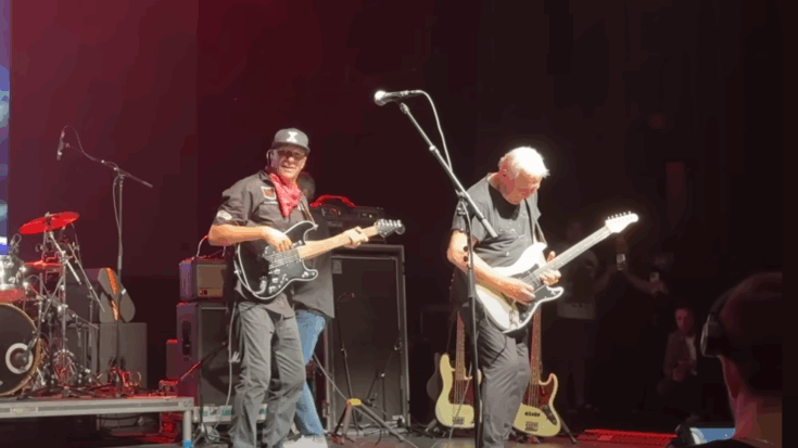 Tom Morello and Alex Lifeson perform together on stage, both playing electric guitars under concert lighting with a full band setup behind them.