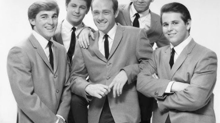 The Beach Boys posing together in matching suits and ties during the early 1960s, smiling at the camera with a clean studio backdrop.