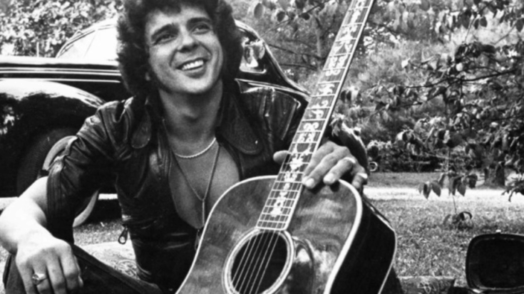 Bobby Whitlock sitting outdoors with an acoustic guitar, smiling, wearing a leather jacket, with a classic car in the background.