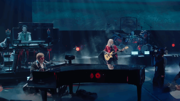 Elton John performs on a grand piano under blue stage lights alongside members of Spinal Tap, including a bassist in a red jacket, a drummer surrounded by a large kit, and a keyboardist dressed in medieval-style clothing, with a film crew capturing the live performance.