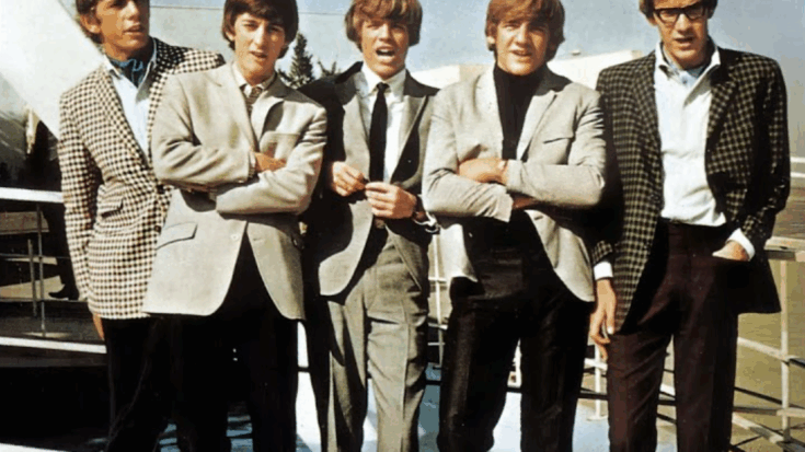 The band Herman's Hermits standing side by side in coordinated suits and jackets during the 1960s, outdoors near a railing.