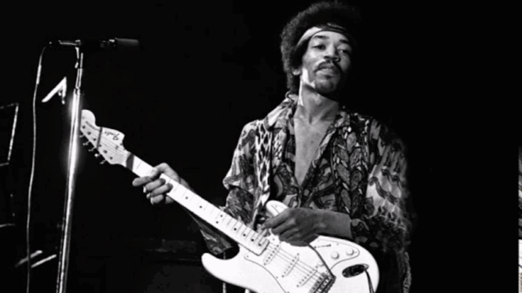 Jimi Hendrix playing electric guitar on stage with a microphone stand beside him.