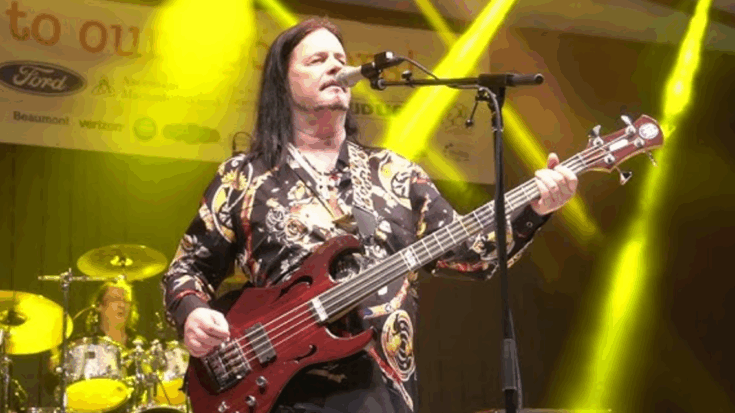 John Payne performing live with Asia Featuring John Payne, playing a red bass guitar and singing into a microphone under bright yellow stage lights.