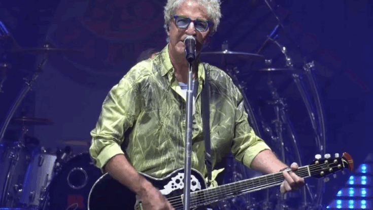 Kevin Cronin performing on stage, playing an acoustic-electric guitar and singing into a microphone, with drums and stage lights in the background.