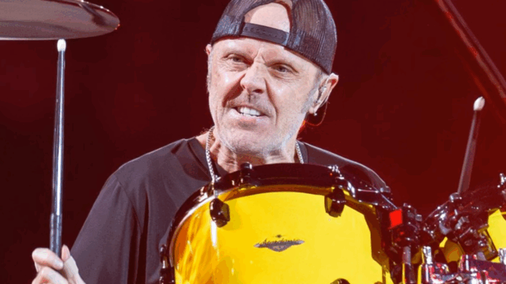 Lars Ulrich performing on stage, playing bright yellow drums while wearing a black shirt and a backwards cap.