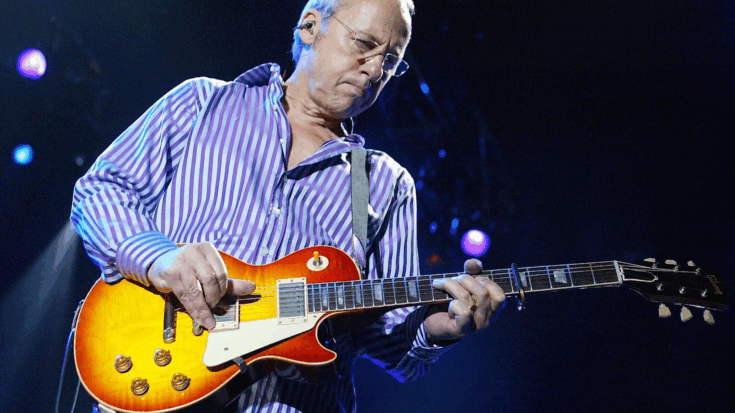 Mark Knopfler wearing a purple striped shirt while playing an electric guitar during a live performance.