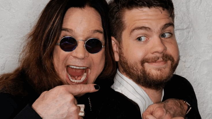 Ozzy Osbourne laughs with his mouth wide open while playfully pointing at his son Jack Osbourne, who looks back at him with a smirk. The two stand back-to-back against a white textured wall, sharing a lighthearted moment.