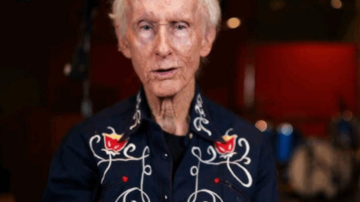 Robby Krieger wearing a dark embroidered shirt with red and white floral designs, speaking in a warmly lit studio with musical instruments in the background.