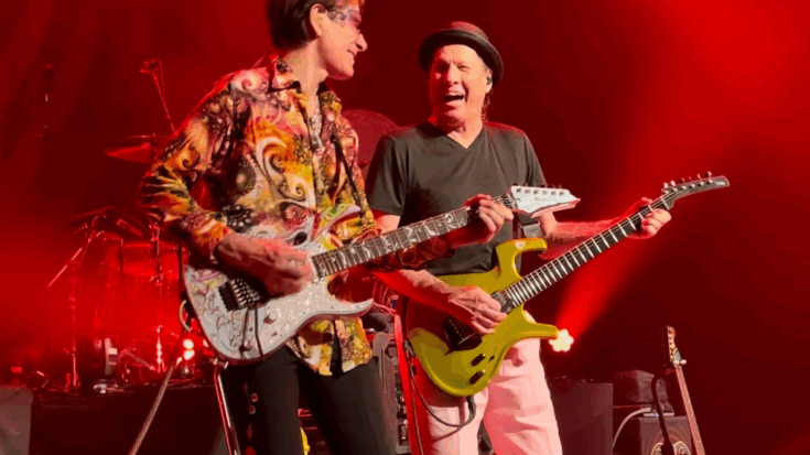 Steve Vai and Adrian Belew performing together on stage under red lighting, both playing electric guitars with expressive energy.