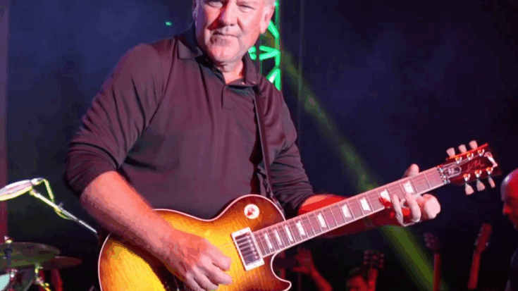 Alex Lifeson performing live on stage playing a sunburst electric guitar under stage lights
