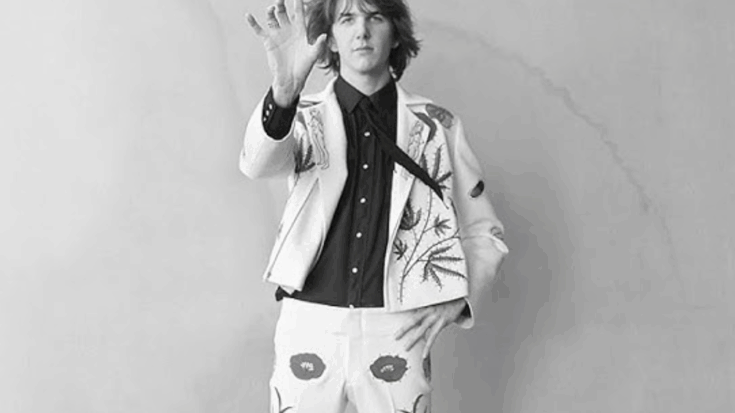 Gram Parsons wearing a white suit decorated with floral embroidery, raising his hand forward against a plain background.