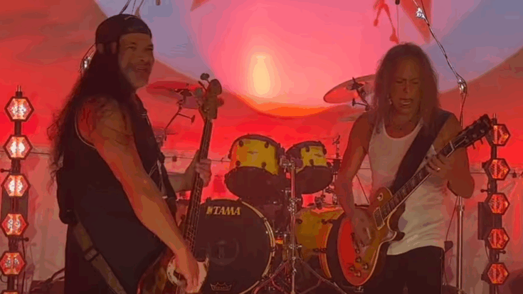 Kirk Hammett and Robert Trujillo performing on stage with red lighting and drums in the background.