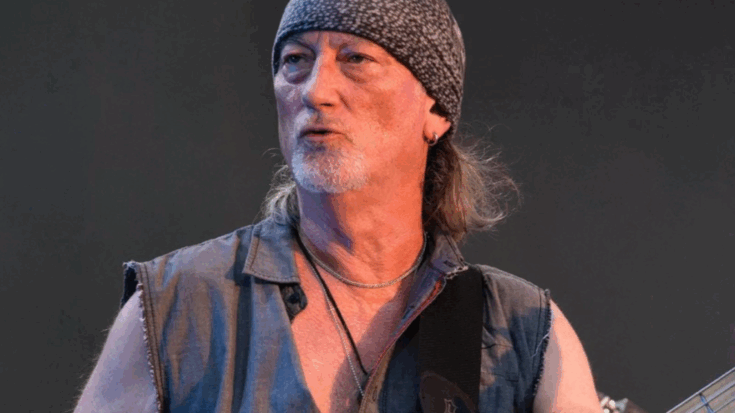 Roger Glover wearing a bandana and sleeveless shirt while performing on stage with his bass guitar.