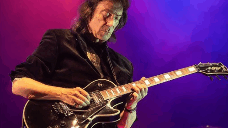 Steve Hackett playing electric guitar live on stage under purple and pink lights.