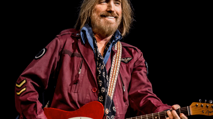 Tom Petty performing on stage with a red guitar, wearing a maroon jacket and smiling under the lights.