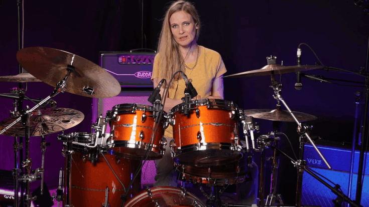 Anika Nilles performing behind an orange drum kit in a studio setup with microphones and amplifiers around her.