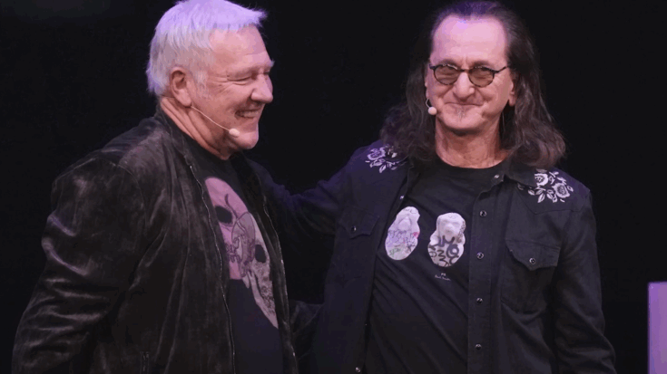 Geddy Lee and Alex Lifeson smiling on stage together during the announcement of Rush’s 2026 reunion tour.