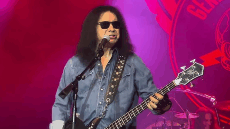 Gene Simmons performing live on stage wearing sunglasses and a denim shirt while playing bass guitar under red stage lights.