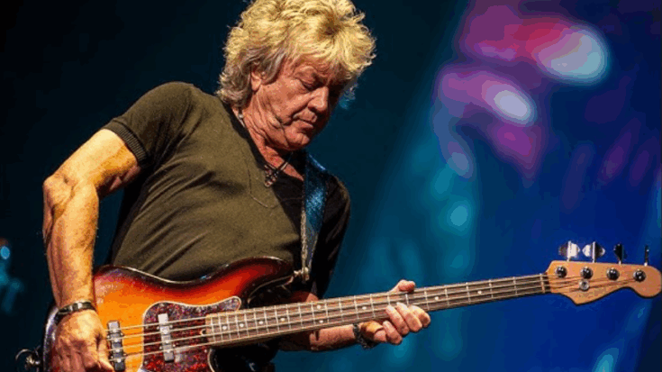 John Lodge performing live on stage, playing a sunburst bass guitar under concert lights.