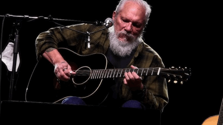 Jorma Kaukonen performing live on stage while playing an acoustic guitar under spotlight.