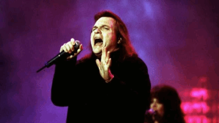 Meat Loaf performing passionately on stage during a live concert, delivering one of his signature powerhouse vocals against a dramatic purple backdrop.