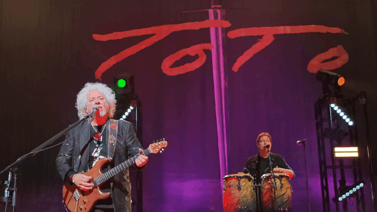 Toto performing live in Paris in 2024, with guitarist Steve Lukather playing under the band’s iconic sword logo backdrop during their performance of “Africa.”