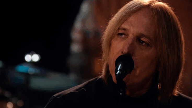 Tom Petty sings into a microphone under warm stage lighting during a live performance.