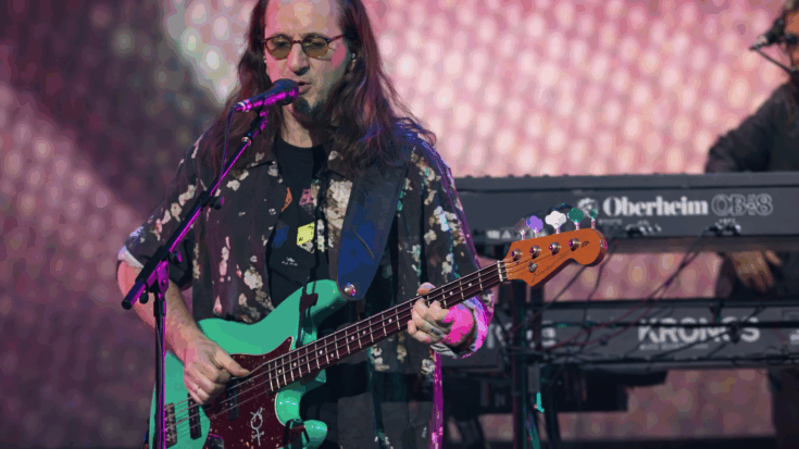 Geddy Lee sings and plays bass onstage under colorful concert lights.