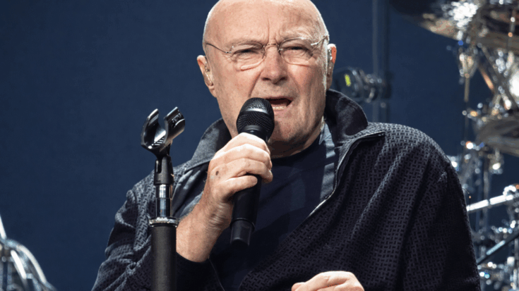 Phil Collins sings into a microphone during a live performance, with his drum kit visible behind him.