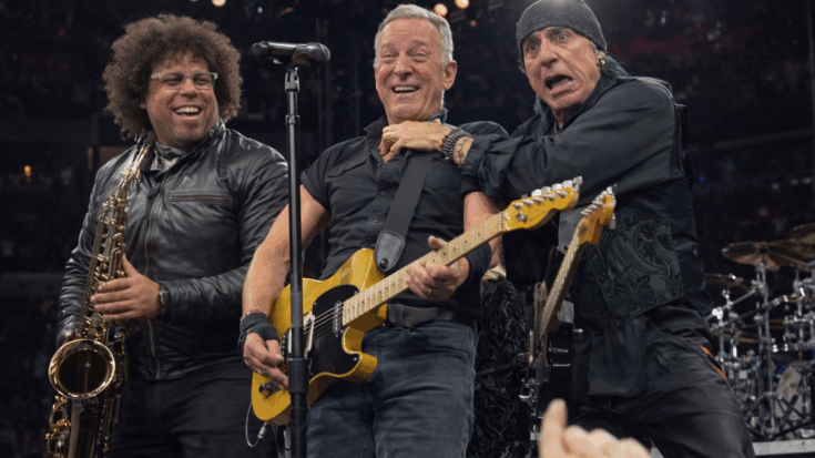 Bruce Springsteen smiles while playing his guitar onstage alongside his bandmates.
