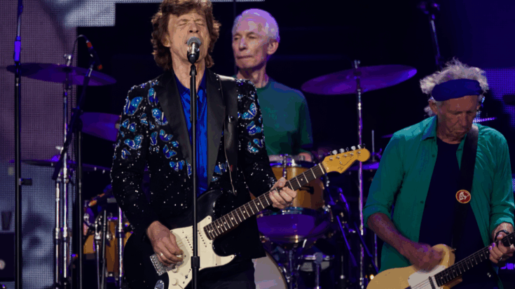Mick Jagger sings at center stage while Charlie Watts plays drums behind him and Keith Richards performs on guitar beside him.