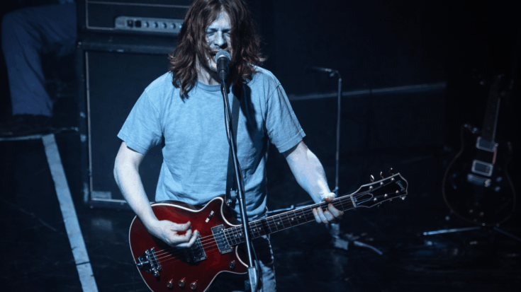 Steve Young performs on stage under blue lighting, singing into a mic while playing a red Les Paul–style electric guitar.
