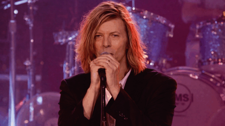 David Bowie performs on stage under warm pink lighting, singing into a microphone with a focused, intense expression.