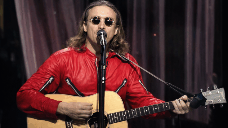 John Lennon performs in a red jacket and sunglasses, singing into a microphone while strumming an acoustic guitar.