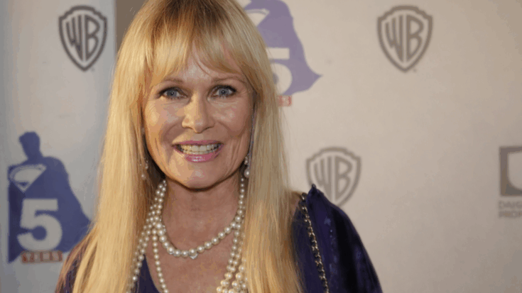 Valerie Perrine smiles warmly at the camera, wearing a purple dress and layered pearl necklace at a red carpet event.