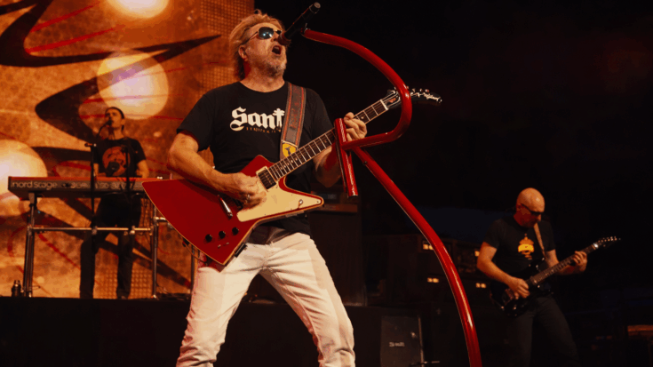 Sammy Hagar rocks the stage with a red Gibson Explorer electric guitar, singing into a curved red mic under bright concert lights.